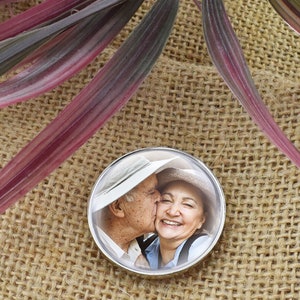 Puede incluir: Un pin redondo plateado con una foto de una pareja mayor besándose. El hombre lleva un sombrero blanco y la mujer un sombrero de paja. La foto está sobre un fondo blanco.