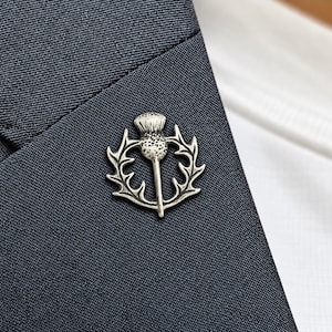 May include: A silver-toned metal brooch shaped like a Scottish thistle, featuring a textured, rounded flower head atop a stem, surrounded by a circular frame of pointed, jagged leaves, pinned onto a dark navy blue textured fabric lapel.