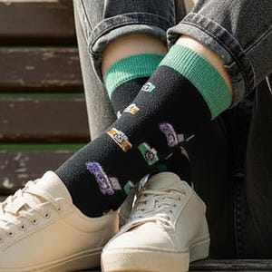 Op de afbeelding: Zwarte sokken met een groene boord en een patroon van camera's, horloges en andere accessoires. De sokken worden gedragen met witte sneakers en een spijkerbroek.