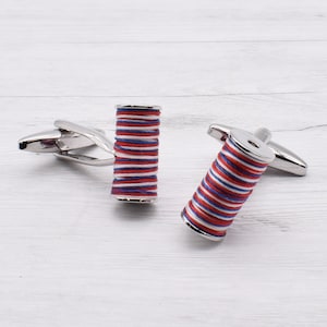 May include: Silver cufflinks featuring a spool design, each wrapped with red, white, and blue thread. The cufflinks are presented on a white wooden surface.