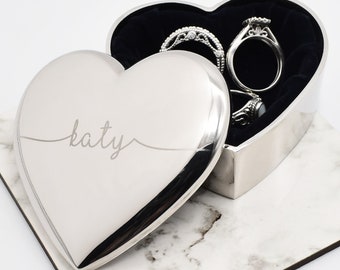 May include: A silver heart-shaped jewelry box with the name "katy" engraved on the lid. The box is open, revealing several rings inside. The interior is lined with black velvet. The box sits on a marble-patterned surface.