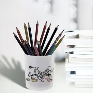May include: White ceramic pencil holder with a colourful design featuring cats and the text "Rebecca is a Crazy Cat Lady". The holder is filled with coloured pencils.