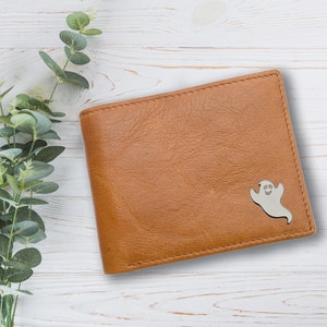 Puede incluir: Una cartera de cuero marrón con un borde cosido y un pequeño diseño de fantasma en la esquina inferior derecha.