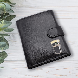 May include: Black leather wallet with a textured finish and a snap closure. The wallet features a gold and black emblem of a pint of stout with the word "Ireland" on it. The wallet is rectangular and has a classic design.