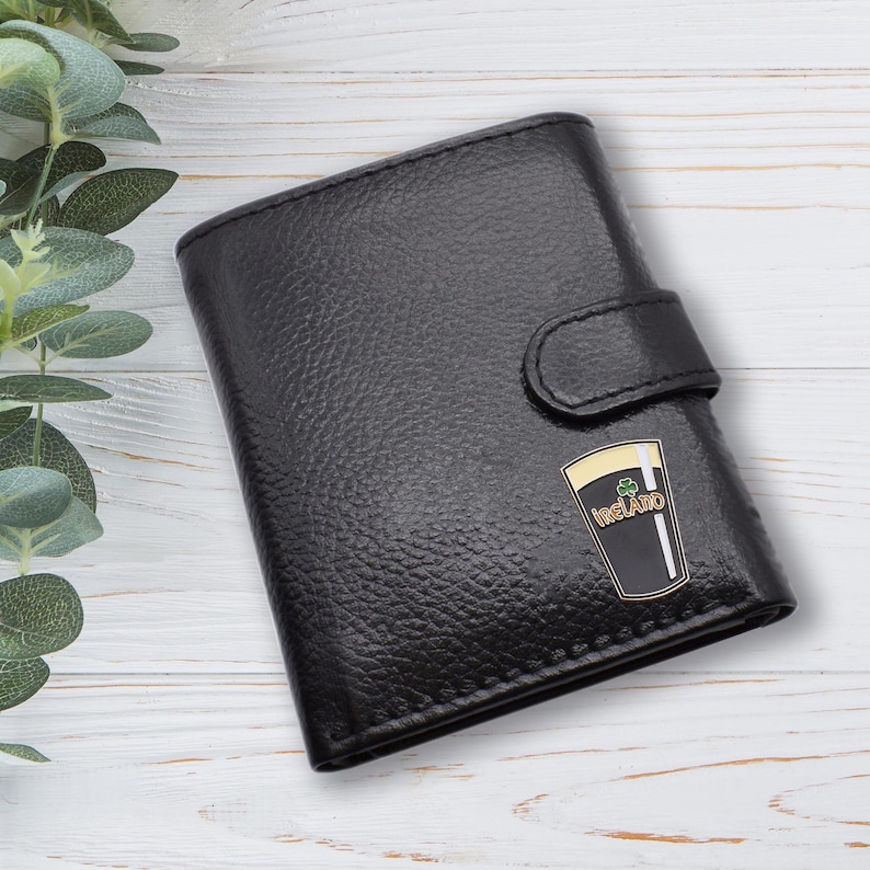 May include: Black leather wallet with a textured finish and a snap closure. The wallet features a gold and black emblem of a pint of stout with the word "Ireland" on it. The wallet is rectangular and has a classic design.