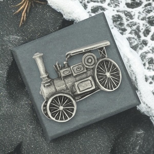 May include: A silver-colored steam engine brooch with detailed wheels and smokestack, resting on a dark gray box. The brooch is a miniature replica of a vintage locomotive. The background includes a textured surface and a hint of white.