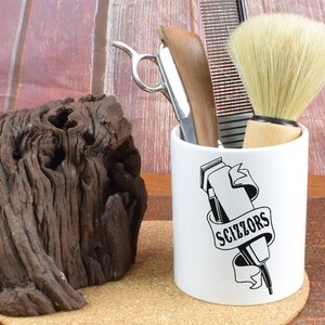May include: White ceramic mug with a black and white illustration of a hair clipper and the text "SCIZZORS". The mug is filled with a straight razor, a comb, and a shaving brush.