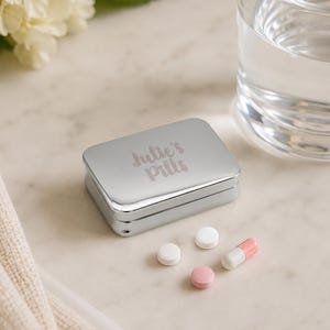 Pode incluir: Uma caixa de comprimidos retangular de metal prateado com o texto gravado "Julie's Pills" na tampa. A caixa está posicionada ao lado de dois comprimidos redondos brancos pequenos, um comprimido redondo rosa pequeno e uma cápsula branca e rosa.