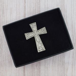 Può includere: Una croce celtica color argento con intricati disegni a nodi, presentata in una scatola nera con interno in velluto. La croce è al centro, mostrando la sua dettagliata lavorazione artigianale. La scatola è rettangolare e poggia su una superficie in legno chiaro.