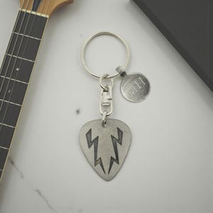 Puede incluir: Un llavero de púa de guitarra plateada con un diseño de rayo. El llavero incluye una pequeña etiqueta redonda con las iniciales "MH". La púa de guitarra está unida a un anillo y un cierre plateados.