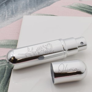 Puede incluir: Un atomizador de perfume plateado con un diseño floral y el nombre "Olivia" grabado en la tapa. El atomizador cilíndrico tiene una boquilla rociadora y una tapa extraíble. El diseño floral está grabado en el cuerpo.