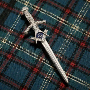 May include: A silver-toned metal kilt pin shaped like a dagger. The handle features intricate textured detailing and a central square and compass emblem with a blue enamel inlay. The blade is smooth and tapers to a point at the bottom end.