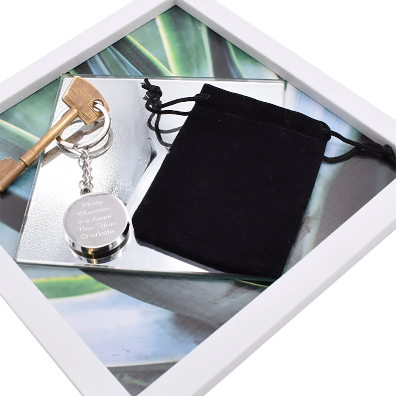 May include: A silver key chain with a round charm that says "Merry Christmas & a Happy New Year Charlotte". The keychain is in a white frame with a green and white background. The keychain is also in a small black velvet bag.