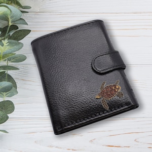 May include: Black leather wallet with a textured finish and a snap closure. A decorative turtle emblem in brown, gold, and tan is affixed to the front. The wallet is rectangular and appears to be closed.