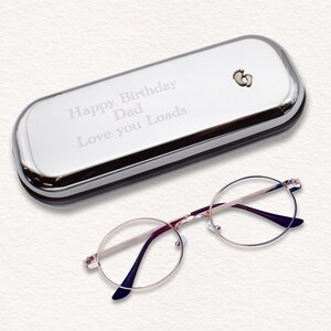May include: A silver eyeglasses case with the text "Happy Birthday Dad Love you Loads" and a small footprint design. Next to the case are round eyeglasses with a gold frame and dark purple arms.