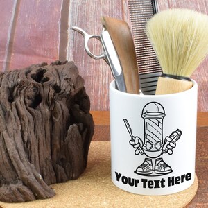 Puede incluir: Taza de cerámica blanca con una ilustración en blanco y negro de un personaje de poste de barbero sosteniendo una navaja y unas tijeras. La taza tiene el texto "Your Text Here" impreso en la parte inferior.