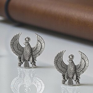 May include: A pair of silver-coloured stud earrings. Each earring features a detailed bird design with outstretched wings, possibly a falcon or eagle. The earrings have an ancient Egyptian aesthetic, with a sun disc above the bird's head. The earrings are on a reflective surface.