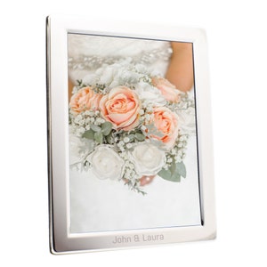 May include: A silver-colored picture frame displaying a wedding bouquet of peach and white roses, with the names "John & Laura" engraved at the bottom. The frame has a clean, modern design.