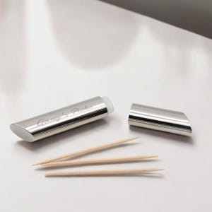 Op de afbeelding: Een gepolijste zilverkleurige metalen tandenstokerhouder met een schuine bovenkant en de gegraveerde tekst "Gary's Picks" aan de zijkant. De houder is geopend met de bijpassende dop rechts ernaast en drie lichte houten tandenstokers liggen ervoor.