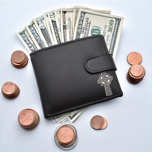 May include: A black leather wallet with a Celtic cross design on the front. The wallet is open and filled with US dollar bills. There are stacks of copper coins scattered around the wallet.