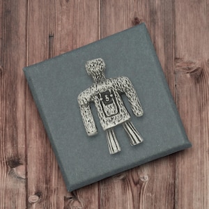 May include: A silver-coloured metal figurine in the shape of a person, lying on a grey box. The figurine has a textured surface and a rectangular design on the chest with the number 5. The box is on a wooden surface.