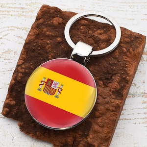 Puede incluir: Un llavero de plata con un charm redondo de la bandera española roja y amarilla. La bandera presenta el escudo de armas español en el centro.