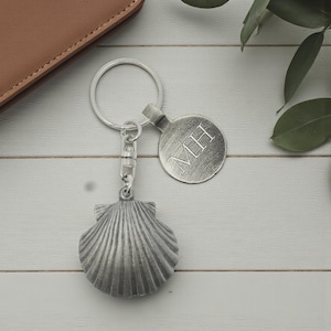 May include: A silver-coloured keyring with a seashell charm and a round tag engraved with the initials "MH". The keyring has a clasp and a key ring. The background is a light wood surface.