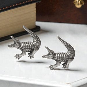 Peut inclure: Deux boutons de manchette argentés en forme d'alligator. Les figurines d'alligator ont la gueule ouverte et des écailles détaillées. Les boutons de manchette sont sur une surface blanche, avec un livre en cuir et une boîte marron en arrière-plan.