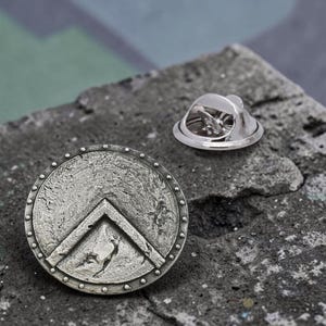 Könnte beinhalten: Eine kreisförmige Anstecknadel aus silberfarbenem Metall mit einem erhabenen, umgedrehten V-förmigen Emblem auf einer texturierten, verwitterten Oberfläche mit genietetem Rand. Die Nadel enthält einen separaten Metallverschluss mit runder Basis.