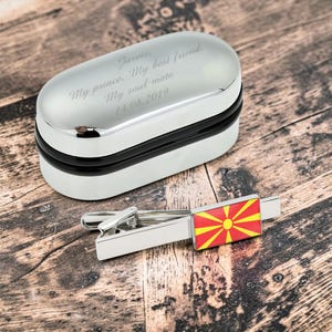 May include: A silver-toned tie clip featuring a rectangular North Macedonia flag emblem in red and yellow, paired with an oval-shaped silver metal storage box engraved with the text: Jarvis, My prince, My best friend, My soul mate, 14.08.2019.