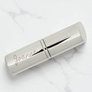 Puede incluir: Un tubo de lápiz labial plateado con el nombre "Joanne" en escritura cursiva. El tubo es cilíndrico con una parte superior e inferior redondeadas. El estuche de lápiz labial está hecho de un metal brillante y reflectante.