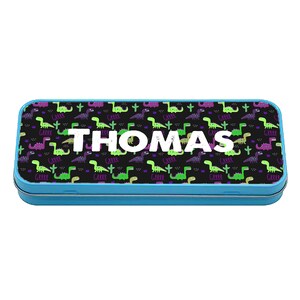 Puede incluir: Un estuche de lápices de metal azul con fondo negro y un patrón repetido de dinosaurios verdes y morados. El nombre "THOMAS" está impreso en letras blancas en la parte delantera del estuche.