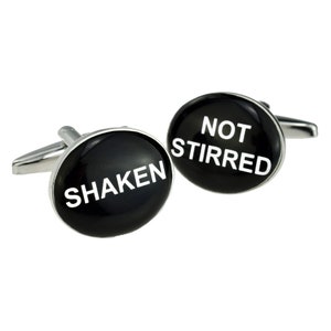 Op de afbeelding: Een paar ovale zilverkleurige manchetknopen met een gepolijst zwart oppervlak. De ene manchetknop bevat de witte tekst SHAKEN, terwijl de andere manchetknop de witte tekst NOT STIRRED bevat. De manchetknopen hebben een standaard metalen sluiting.