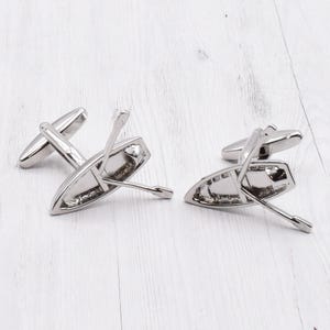 May include: A pair of silver cufflinks featuring a small boat with oars. The boat is tilted to the side, as if it is floating on water.