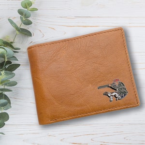May include: A brown leather wallet with a stitched edge and a silver metal pin of a hunter with a dog.