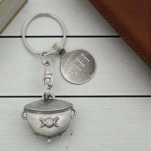 May include: A silver-colored keychain featuring a cauldron charm with a triple moon symbol and a round tag engraved with "MH". The keychain is attached to a large metal ring.