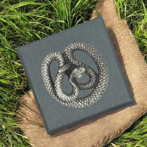 May include: A square, dark grey canvas print featuring a detailed, grayscale illustration of a coiled snake. The artwork is set against a backdrop of green grass and a burlap-textured surface.