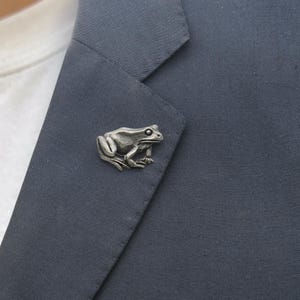 May include: A silver-toned metal frog lapel pin featuring a detailed, textured surface with defined eyes and limbs, attached to the dark charcoal grey fabric of a blazer lapel, showcasing a realistic amphibian design in a metallic finish.