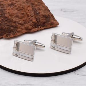 May include: A pair of silver cufflinks with a white inlay. The cufflinks are rectangular in shape and have a polished finish.