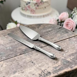 May include: A silver-toned cake knife and server set with polished metal blades and ornate handles. The knife blade features an engraving that reads Congratulations Mr. & Mrs. Adams. The utensils have a smooth, reflective finish and decorative detailing at the base of the handles.