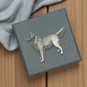 Puede incluir: Una figura de perro plateado se muestra sobre una caja gris oscuro. El perro está de pie con la cola levantada. La caja está sobre una superficie de madera, con una tela azul claro en el fondo.