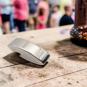 May include: A shiny, silver bottle opener with the letters "MEH" engraved on top. The opener is resting on a weathered wooden surface, with a blurred background of people and a beer bottle.
