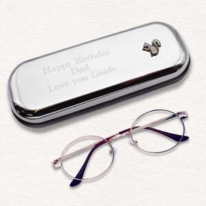 Może przedstawiać: Srebrne etui na okulary z wygrawerowanym napisem "Happy Birthday Dad Love you Loads" i małym wzorem wiewiórki. Etui jest otwarte, a obok leży para okrągłych okularów do czytania w złotej oprawie.