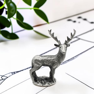May include: A small, silver-coloured metal figurine of a standing stag with detailed antlers and textured fur. The deer is depicted in a side profile view, standing on a circular base, showcasing a metallic finish and realistic anatomical features.