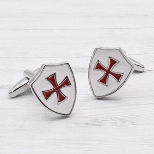 May include: A pair of silver cufflinks with a white enamel shield design. The shield features a red cross in the center.