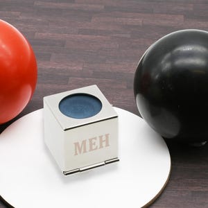 May include: A white, square box with a silver top and hinge, engraved with the letters "MEH". The box has a circular blue interior. A red and a black sphere are on either side of the box, on a white surface.