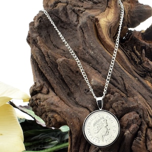 Puede incluir: Un collar de cadena de plata con un colgante de moneda de plata que presenta un perfil de la reina Isabel II y el año 2002. El collar se muestra sobre una pieza de madera desgastada.