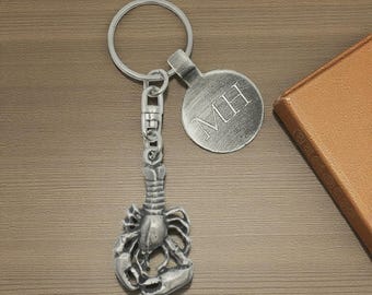 May include: A silver-colored lobster keychain with a round tag engraved with the initials "MH". The lobster charm is detailed and attached to a key ring and clasp. The keychain is on a wooden surface.