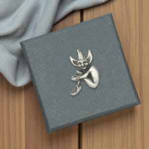 Puede incluir: Figura plateada de un elfo sentado con orejas puntiagudas y un sombrero, sobre una caja gris oscuro. El elfo se abraza las piernas, y la caja está sobre una superficie de madera. Un paño azul claro está drapeado en el fondo.