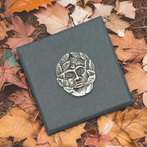 May include: A silver brooch with a face design encircled by leaves, resting on a dark grey box. The background shows autumnal leaves in brown and orange hues, creating a seasonal aesthetic.